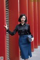 A woman leaning against a red wall wearing a black shirt and blue skirt.