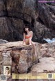 A woman in a bikini sitting on a rock by the ocean.