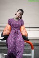 A woman in a purple dress and orange jacket leaning against a railing.